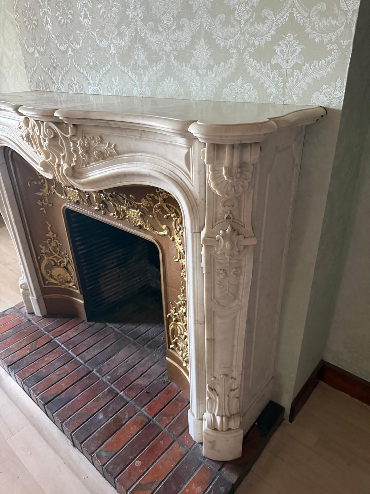 Cheminée de style Louis XV en marbre blanc vue de trois quarts : jambage sculpté d'une coquille et de feuillages, tablette moulurée et intérieur de fonte à décor doré